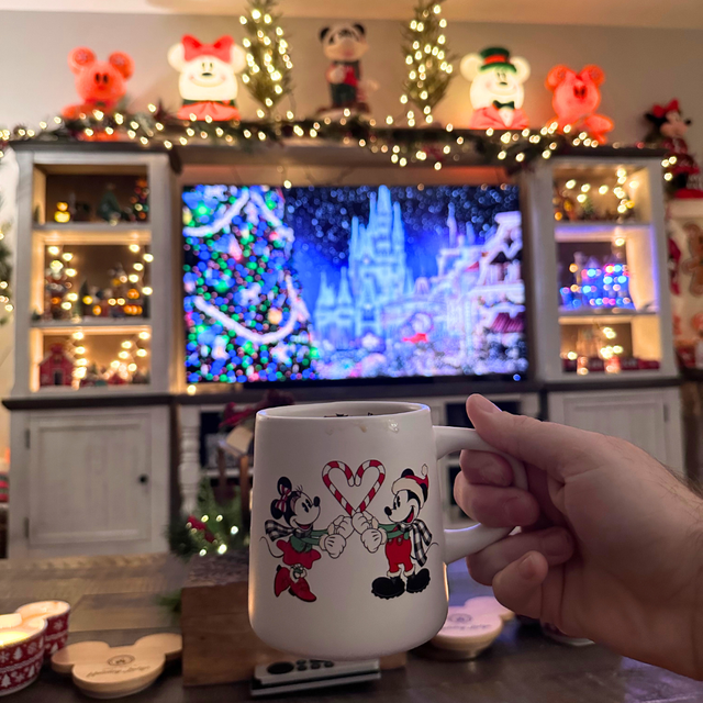 A hand holding a mug of hot cocoa in a living room glowing with Christmas lights.