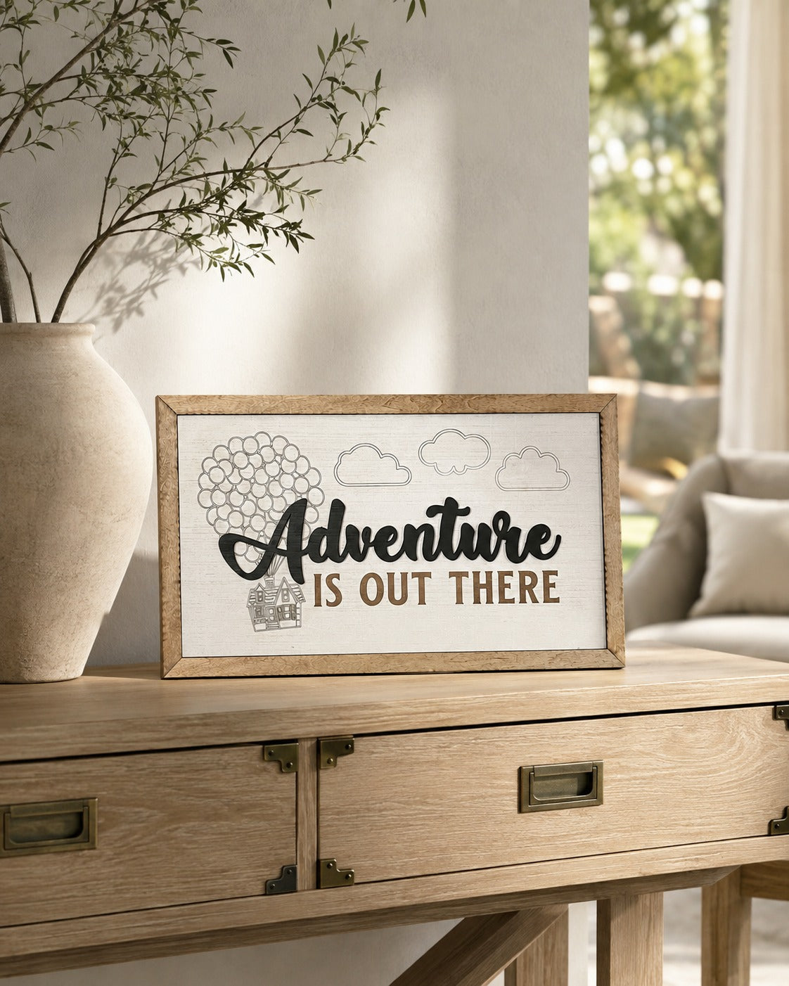 Adventure Is Out There balloon house framed wood sign styled on a light oak console in a sunlit conservatory