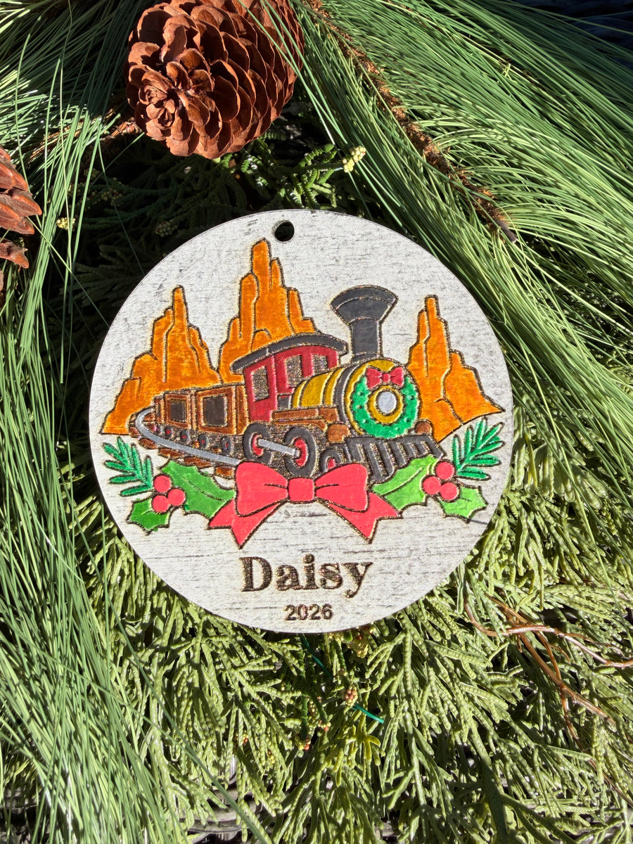 Finished hand-painted Big Thunder Mountain Christmas Ornament on evergreen greenery. Name and year shown are sample personalization.