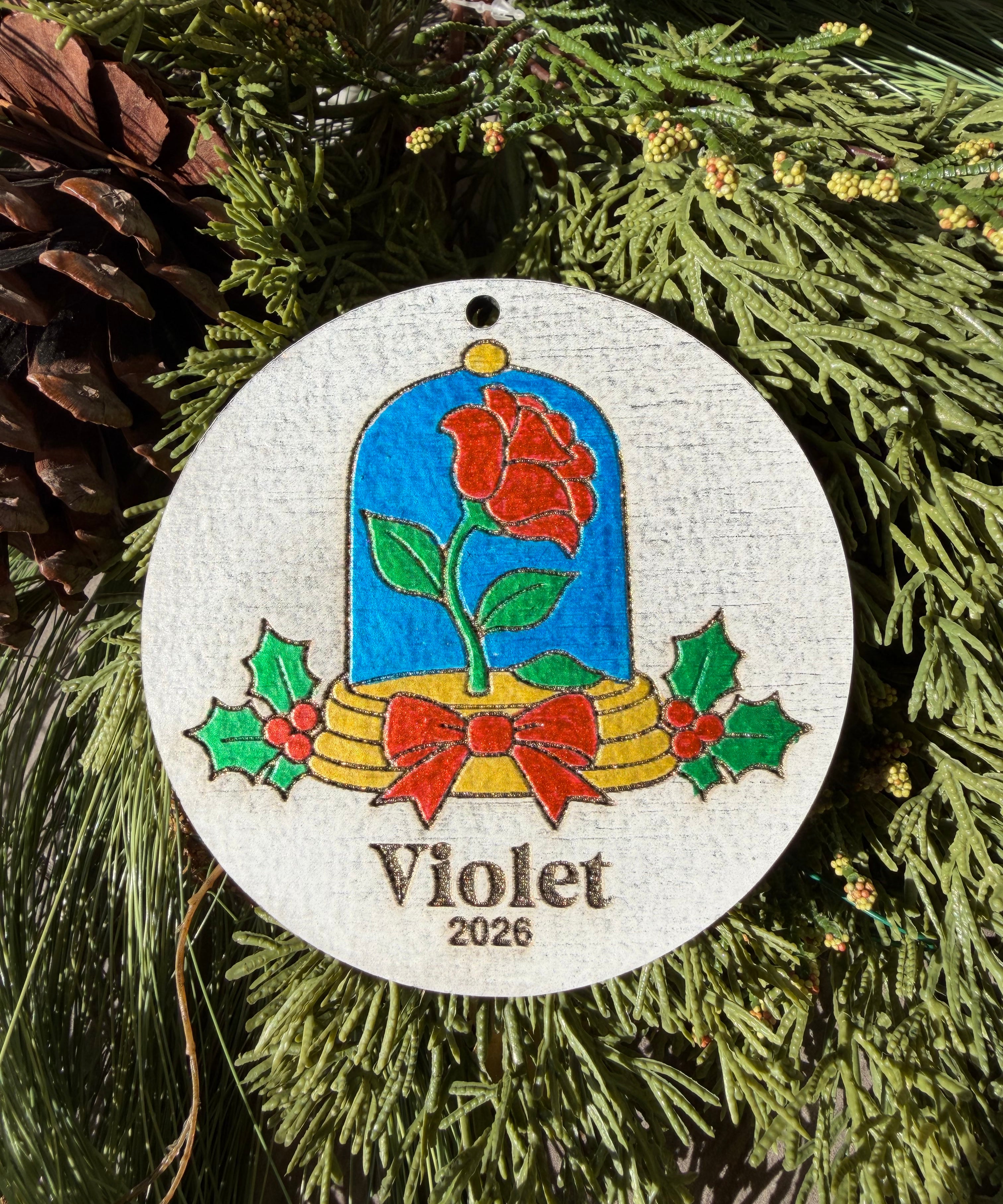 Finished hand-painted Enchanted Rose ornament on evergreen greenery. Violet and 2026 show sample personalization; standard comes without a name or year.