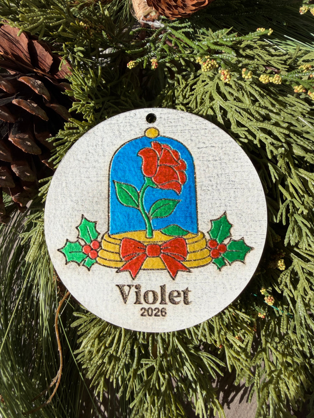 Finished hand-painted Enchanted Rose ornament on evergreen greenery. Violet and 2026 show sample personalization; standard comes without a name or year.