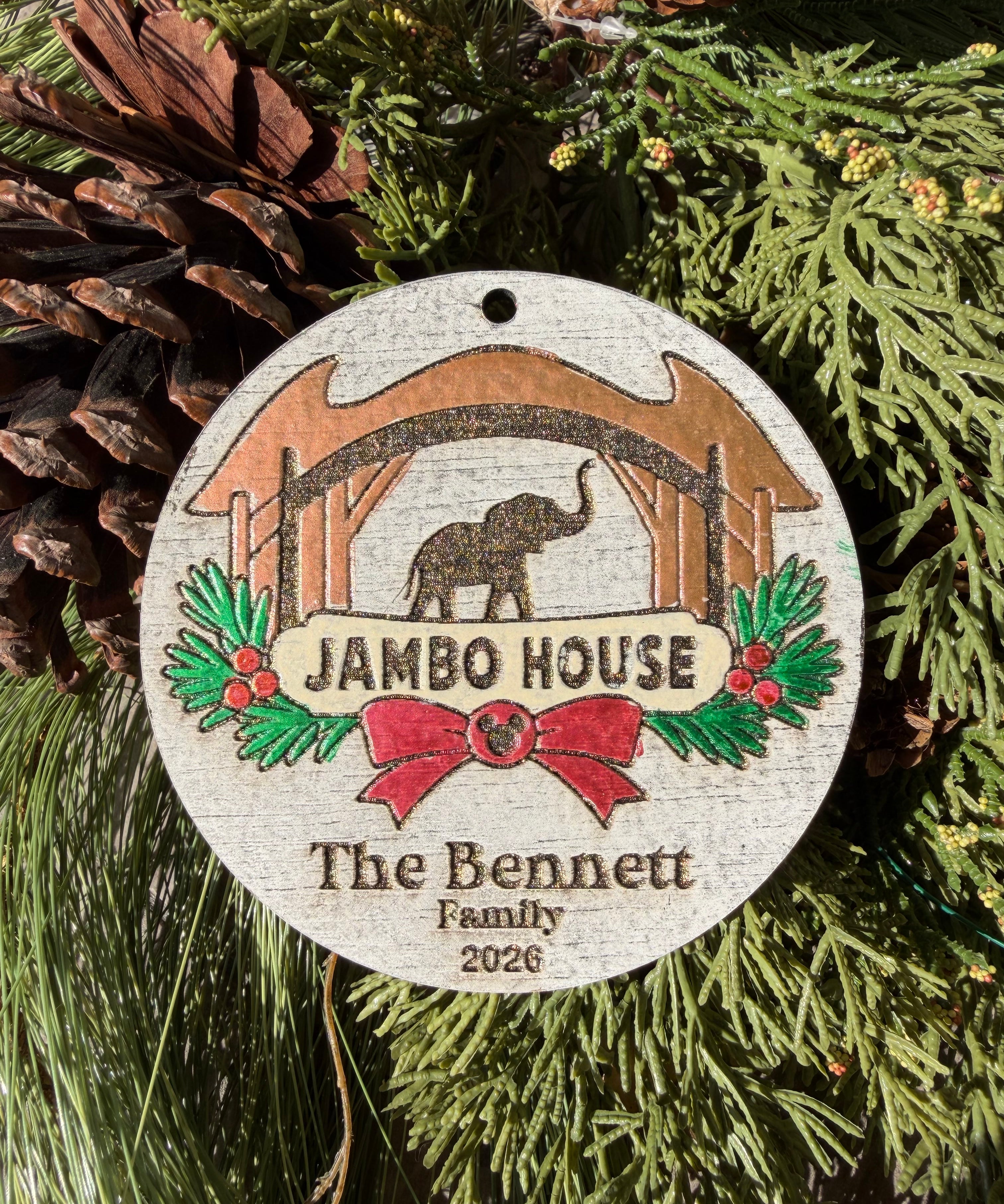 Finished hand-painted Jambo House Christmas Ornament on evergreen greenery. Personal name and year shown are sample personalization.