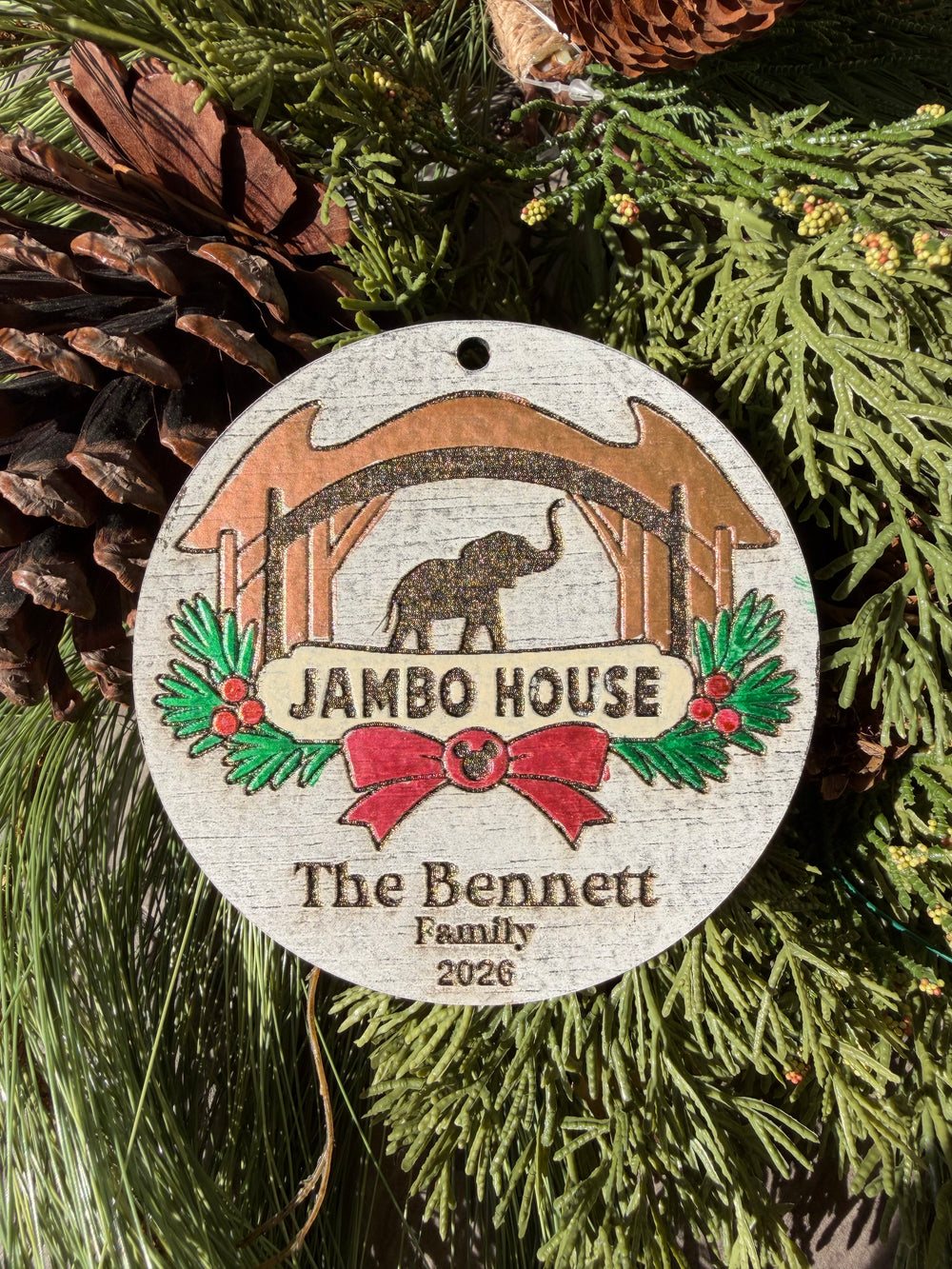 Finished hand-painted Jambo House Christmas Ornament on evergreen greenery. Personal name and year shown are sample personalization.