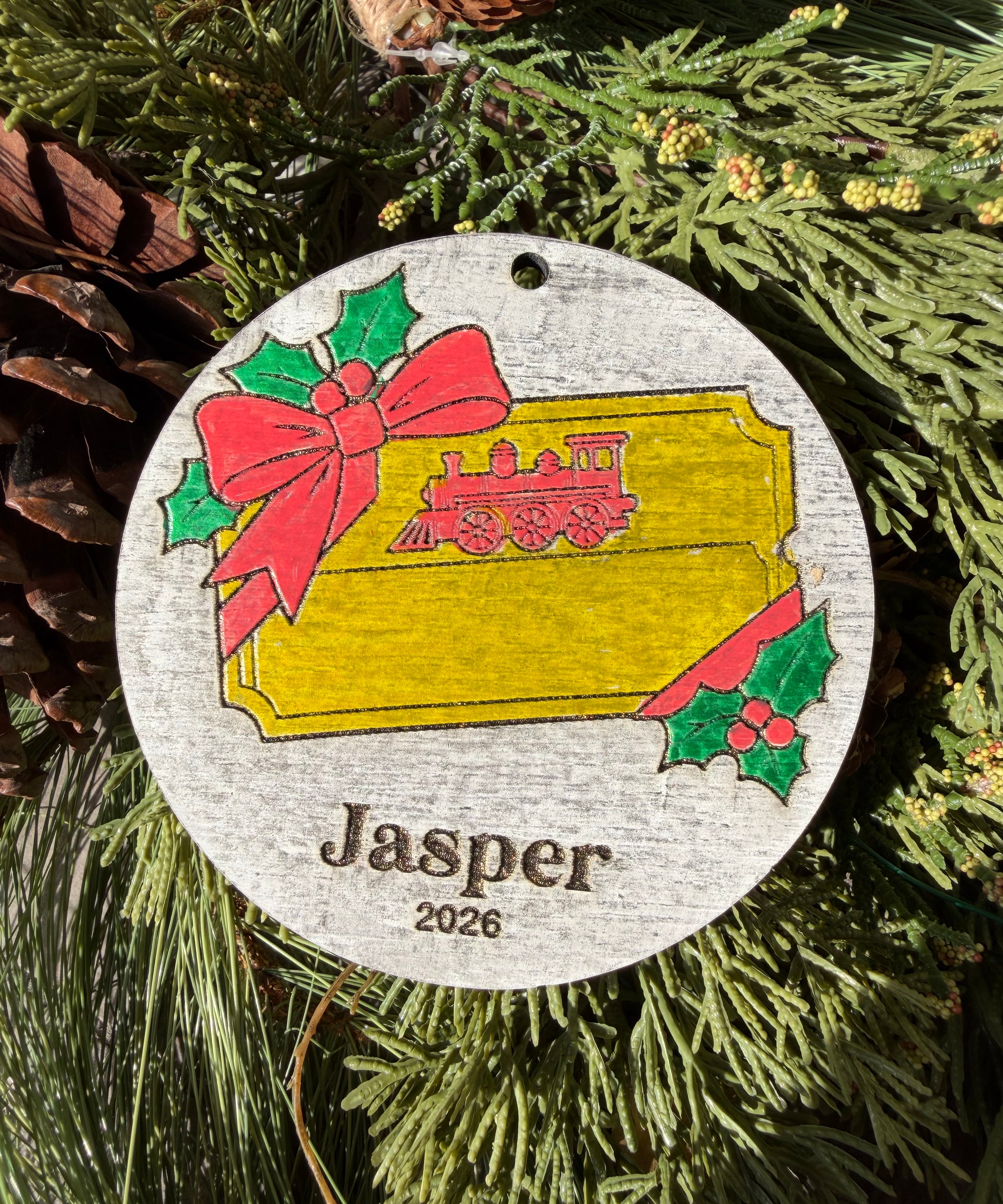 Finished hand-painted Main Street Railroad Ticket Christmas Ornament on evergreen greenery. Personal name and year shown are sample personalization.