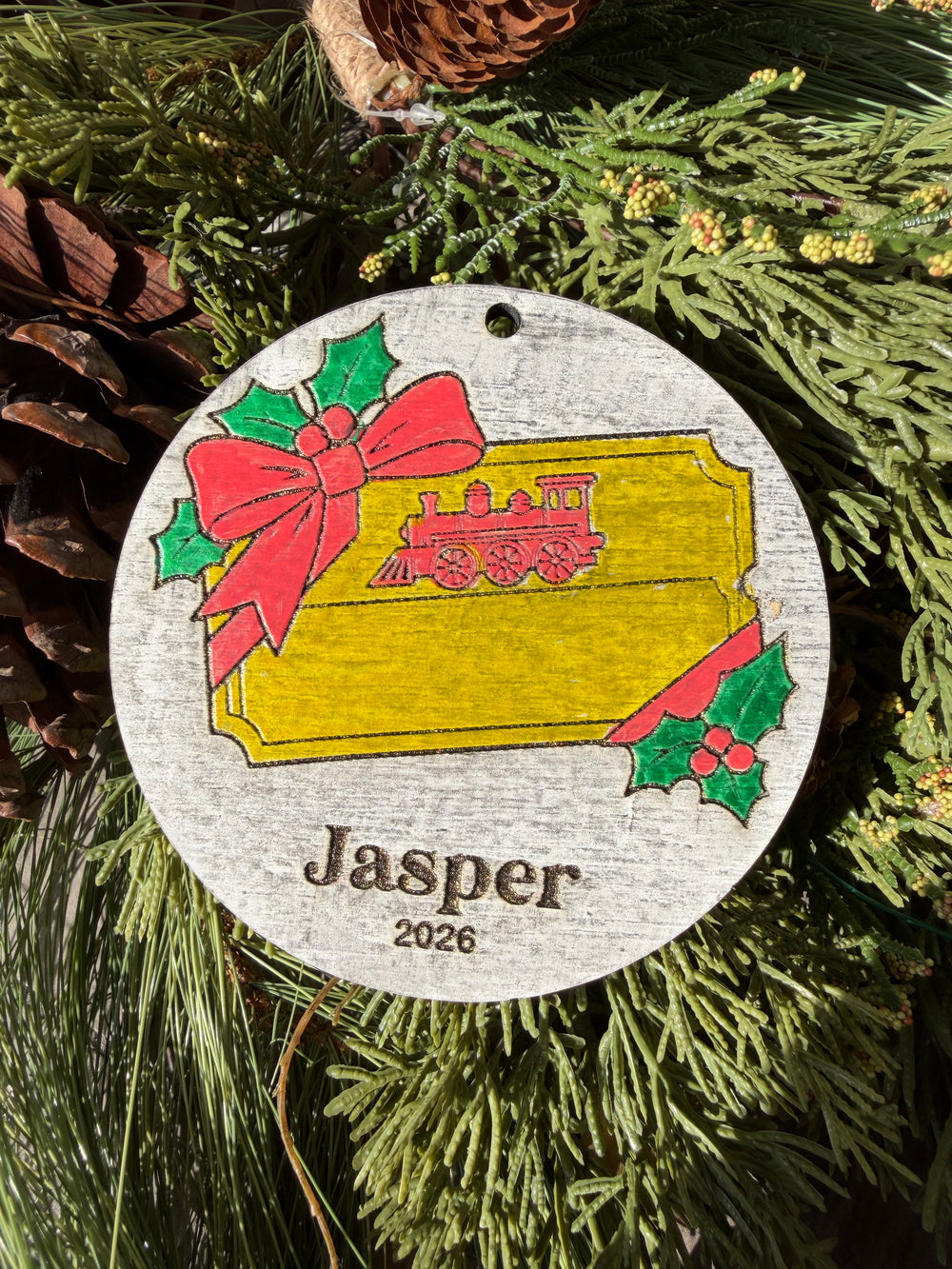 Finished hand-painted Main Street Railroad Ticket Christmas Ornament on evergreen greenery. Personal name and year shown are sample personalization.