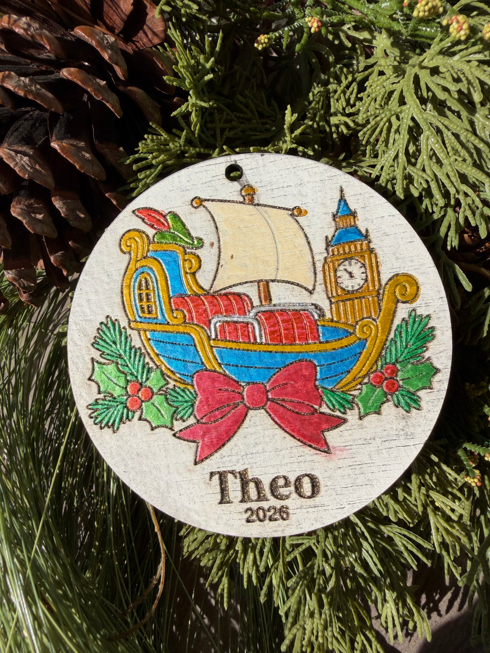 Finished hand-painted Neverland Flying Ship Christmas Ornament on evergreen greenery. Personal name and year shown are sample personalization.