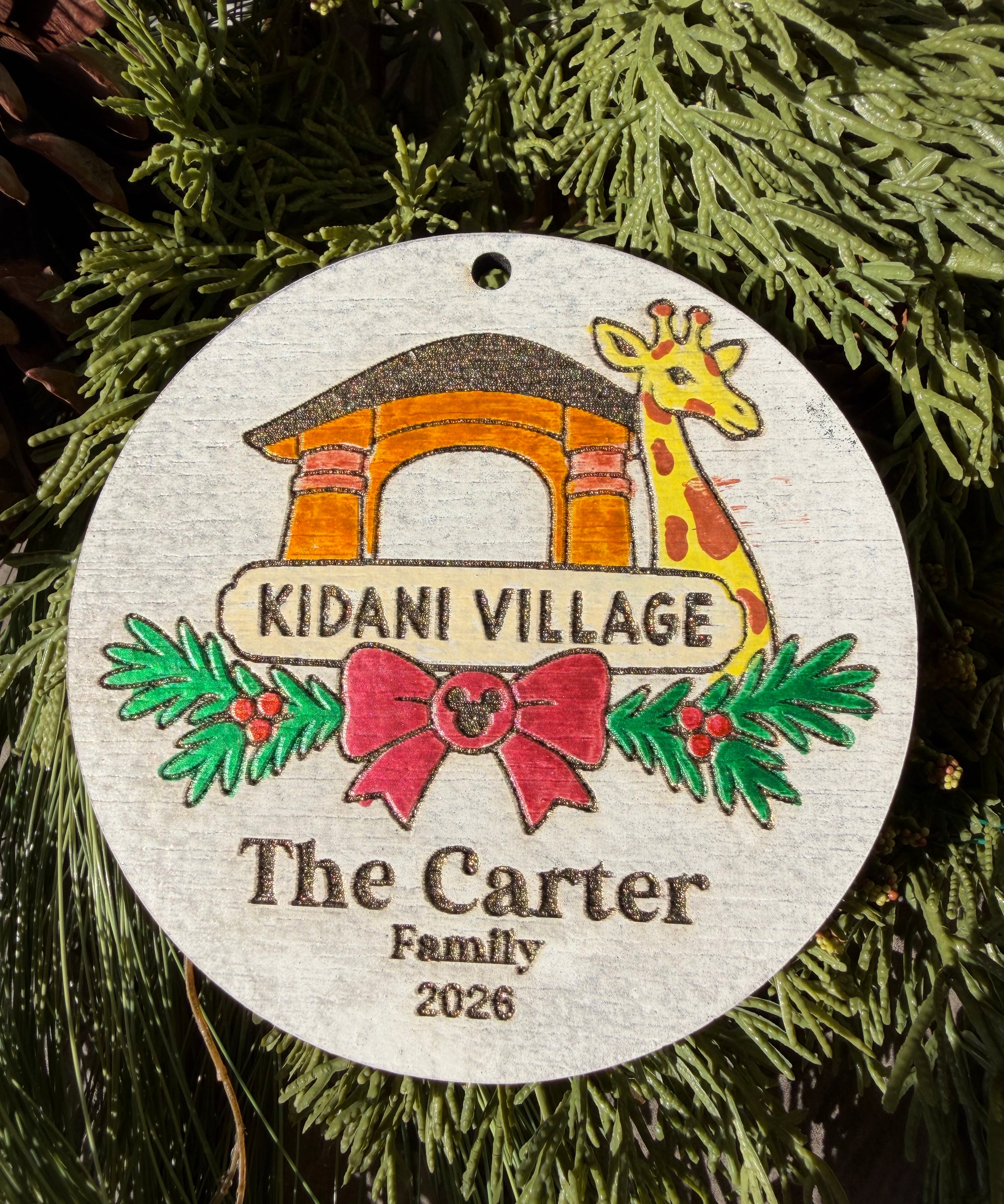 Finished hand-painted Kidani Village Christmas Ornament on evergreen greenery. Personal name and year shown are sample personalization.