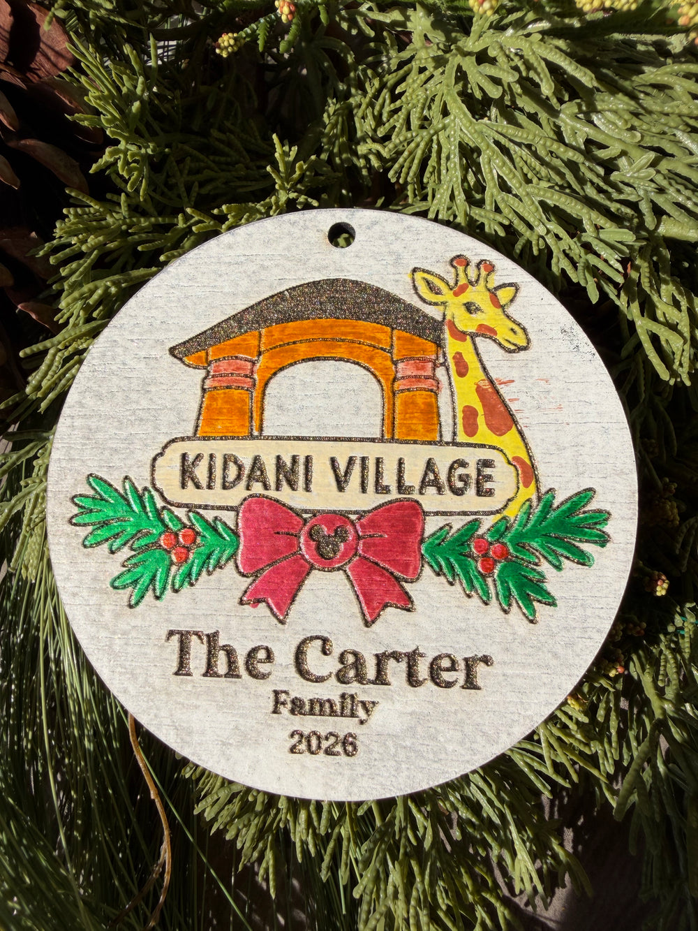 Finished hand-painted Kidani Village Christmas Ornament on evergreen greenery. Personal name and year shown are sample personalization.