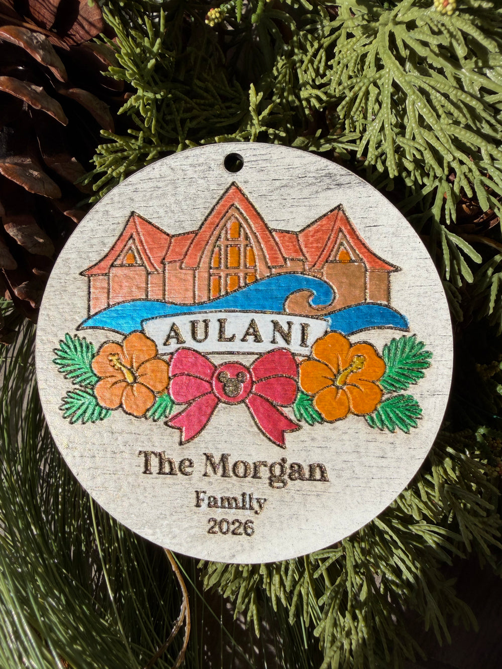 Finished hand-painted Aulani Christmas Ornament on evergreen greenery. Personal name and year shown are sample personalization.