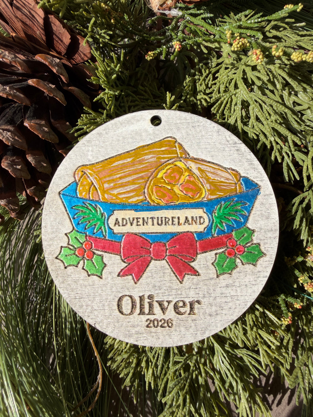 Finished hand-painted Adventureland Holiday Entrance on evergreen greenery. Personal name and year shown are sample personalization.