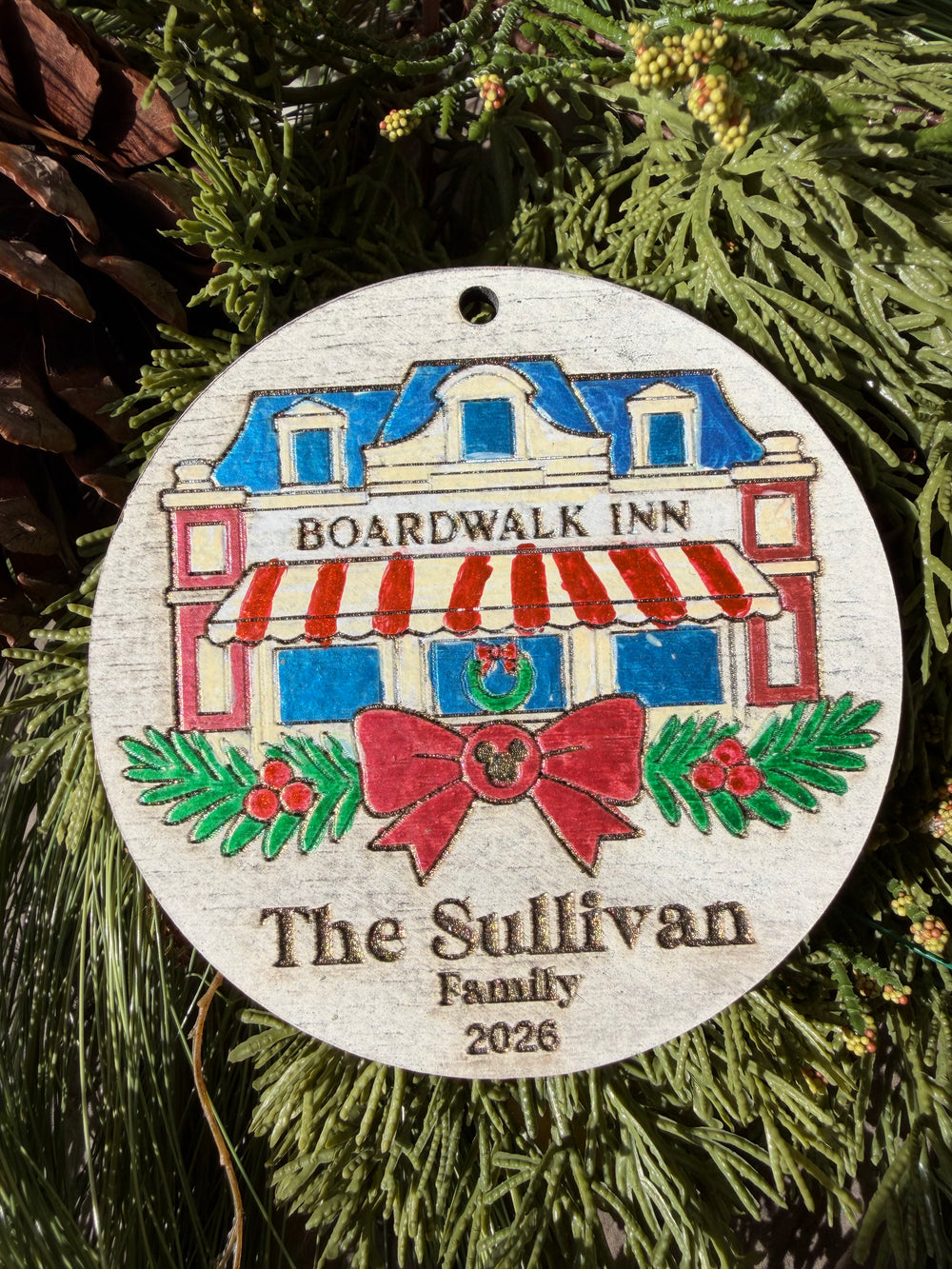 Finished hand-painted BoardWalk Inn Christmas Ornament on evergreen greenery. Personal name and year shown are sample personalization.