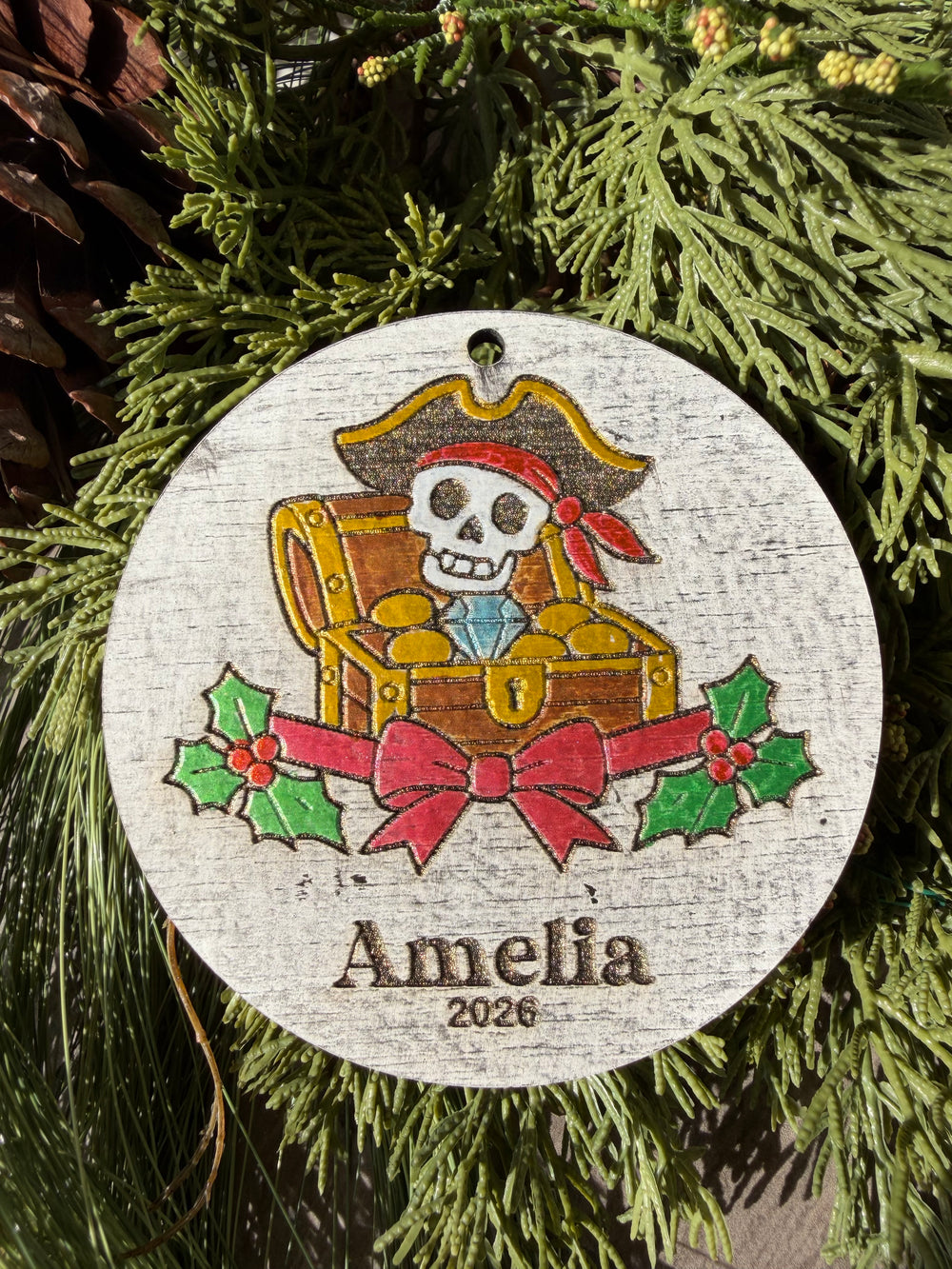 Finished hand-painted Pirate Treasure Christmas Ornament on evergreen greenery. Personal name and year shown are sample personalization.