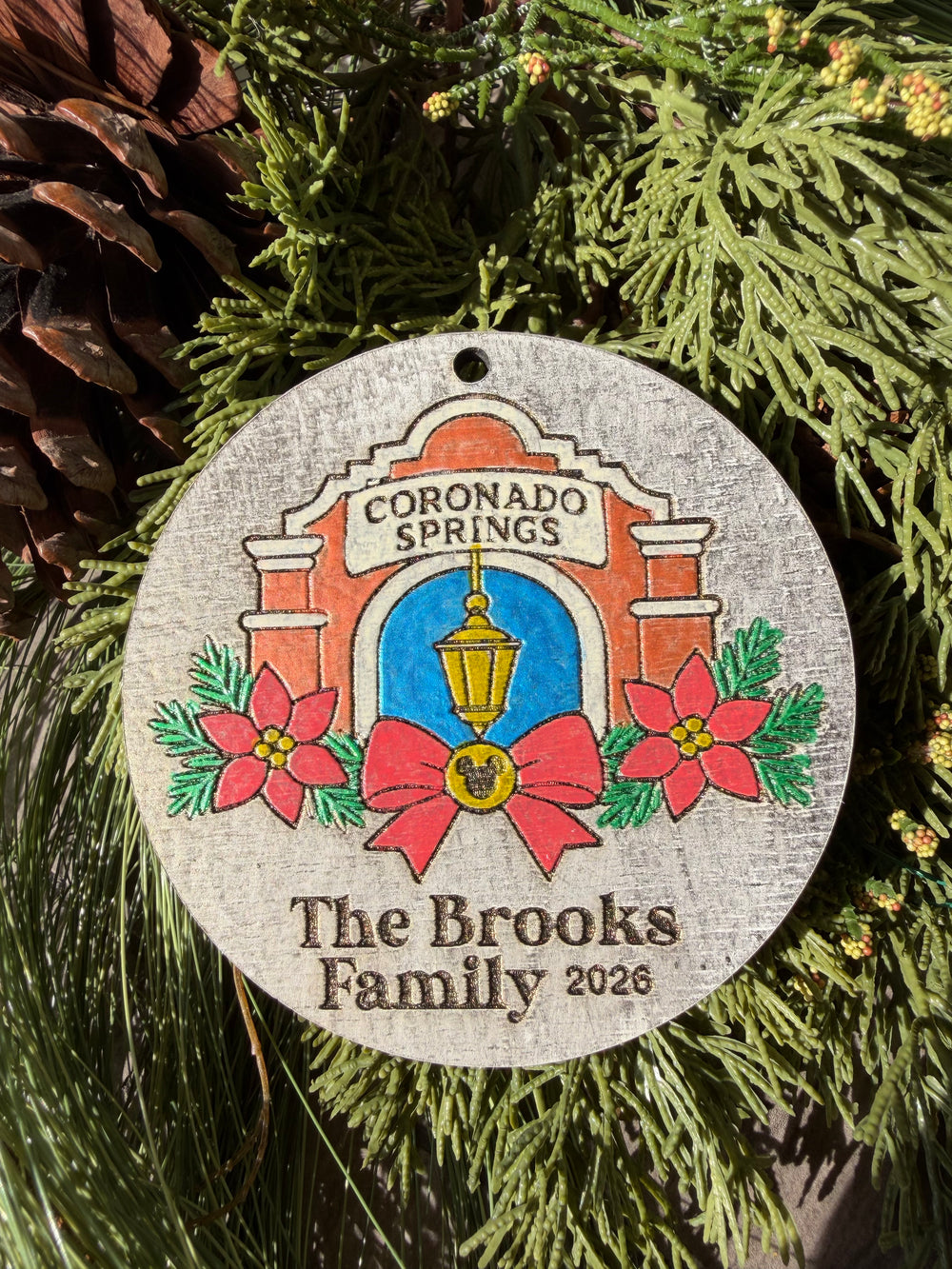 Finished hand-painted Coronado Springs Christmas Ornament on evergreen greenery. Personal name and year shown are sample personalization.