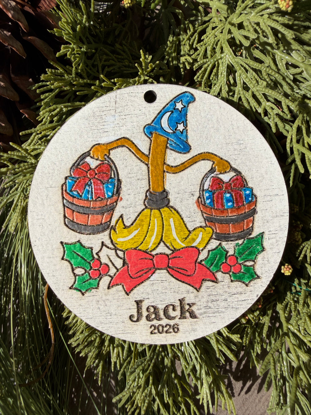 Finished hand-painted Enchanted Broom & Gifts Christmas Ornament on evergreen greenery. Personal name and year shown are sample personalization.