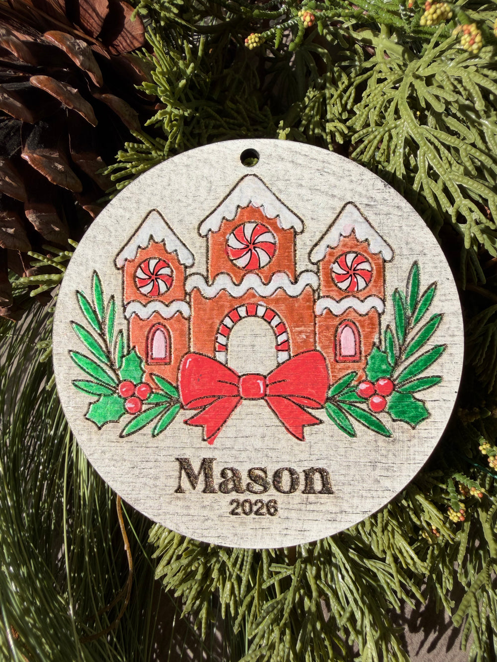 Finished hand-painted Gingerbread Castle Christmas Ornament on evergreen greenery. Personal name and year shown are sample personalization.