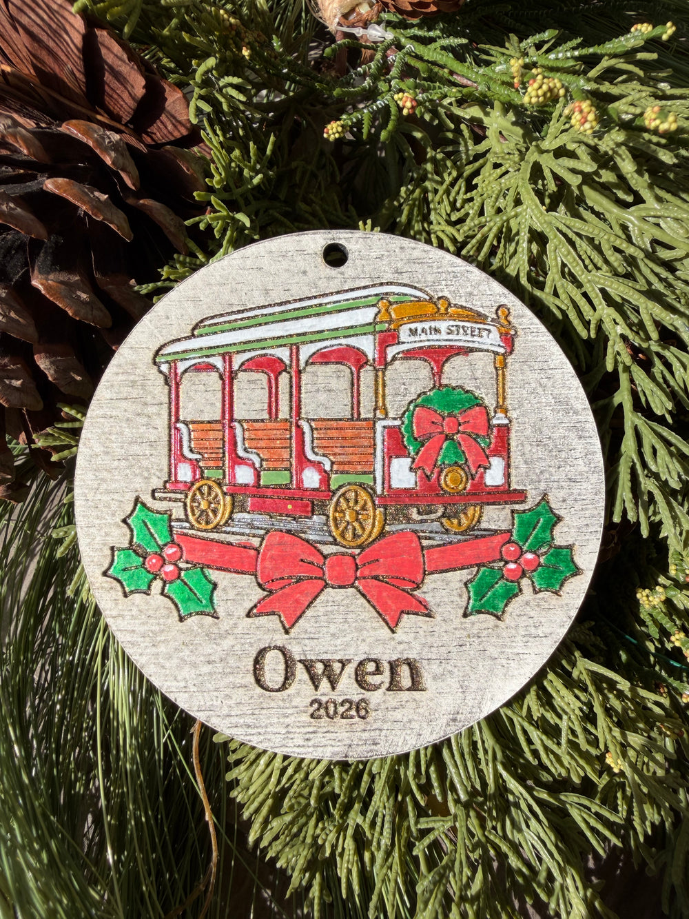 Finished hand-painted Main Street Trolley Christmas Ornament on evergreen greenery. Personal name and year shown are sample personalization.
