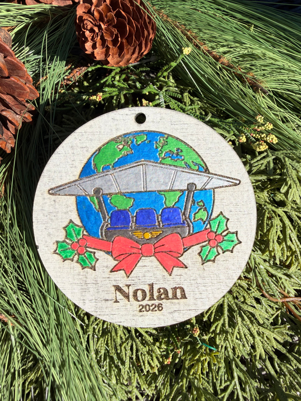 Finished hand-painted Soarin’ Around the World Christmas Ornament on evergreen greenery. Name and year shown are sample personalization.