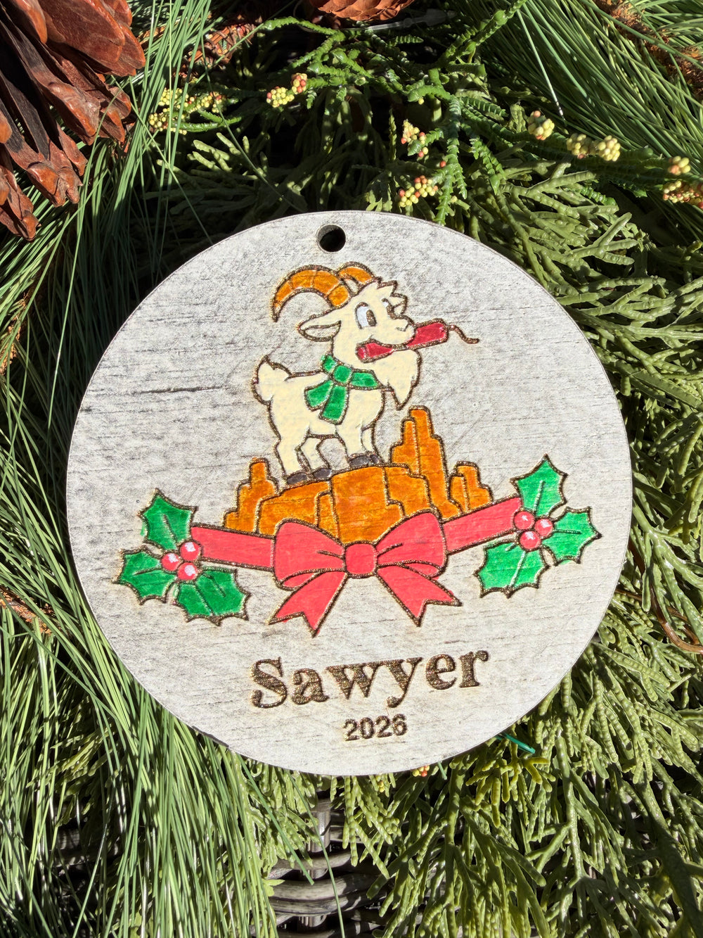 Finished hand-painted Thunder Mountain Goat Christmas Ornament on evergreen greenery. Name and year shown are sample personalization.