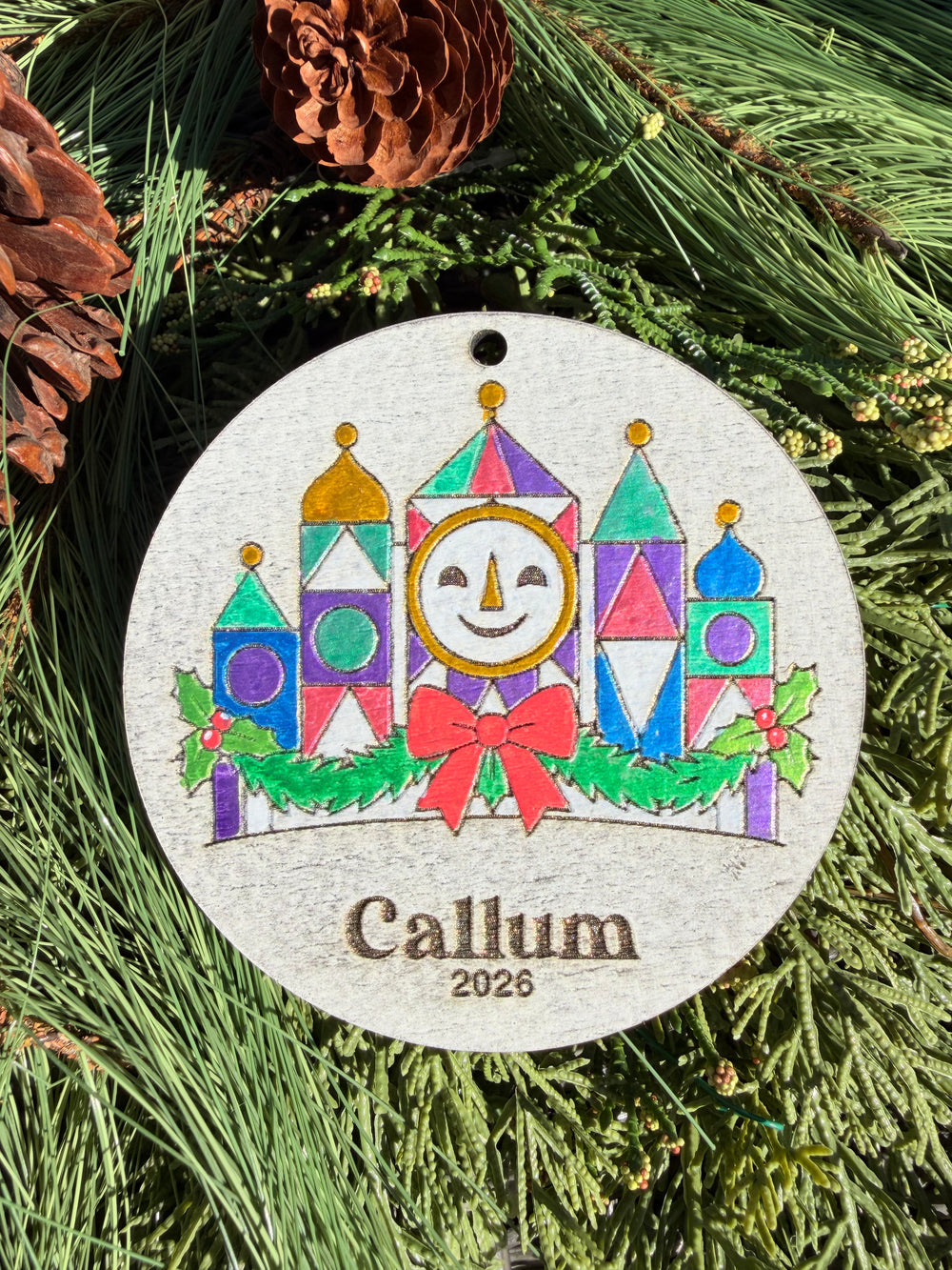 Finished hand-painted Small World Holiday Clock Christmas Ornament on evergreen greenery. Name and year shown are sample personalization.
