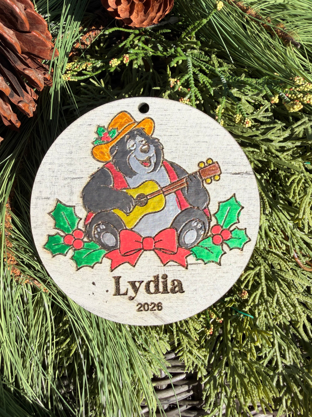Finished hand-painted Country Bear Christmas Ornament on evergreen greenery. Name and year shown are sample personalization.