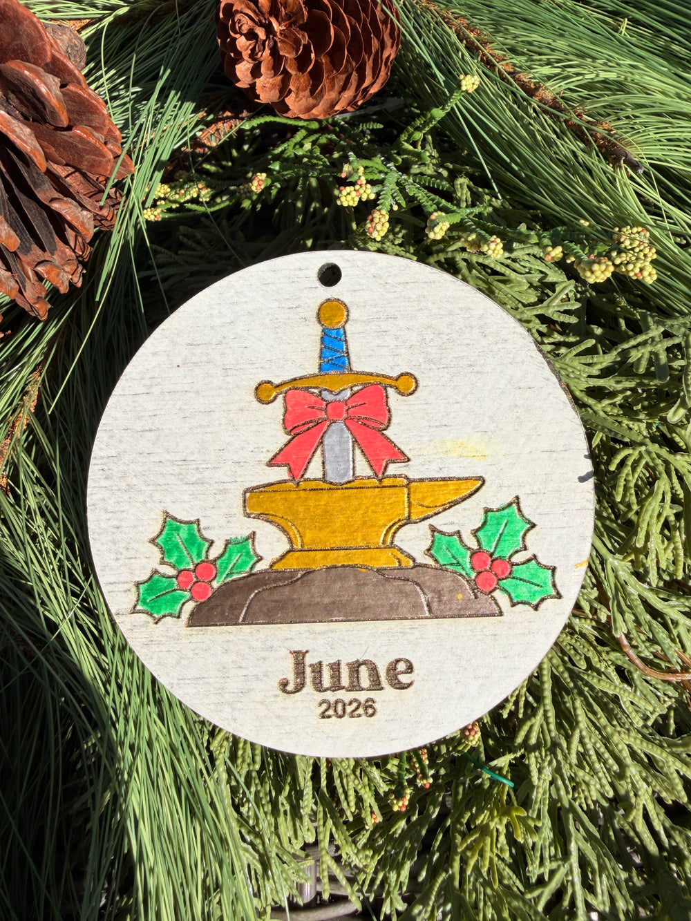 Finished hand-painted Sword in the Stone Christmas Ornament on evergreen greenery. Name and year shown are sample personalization.