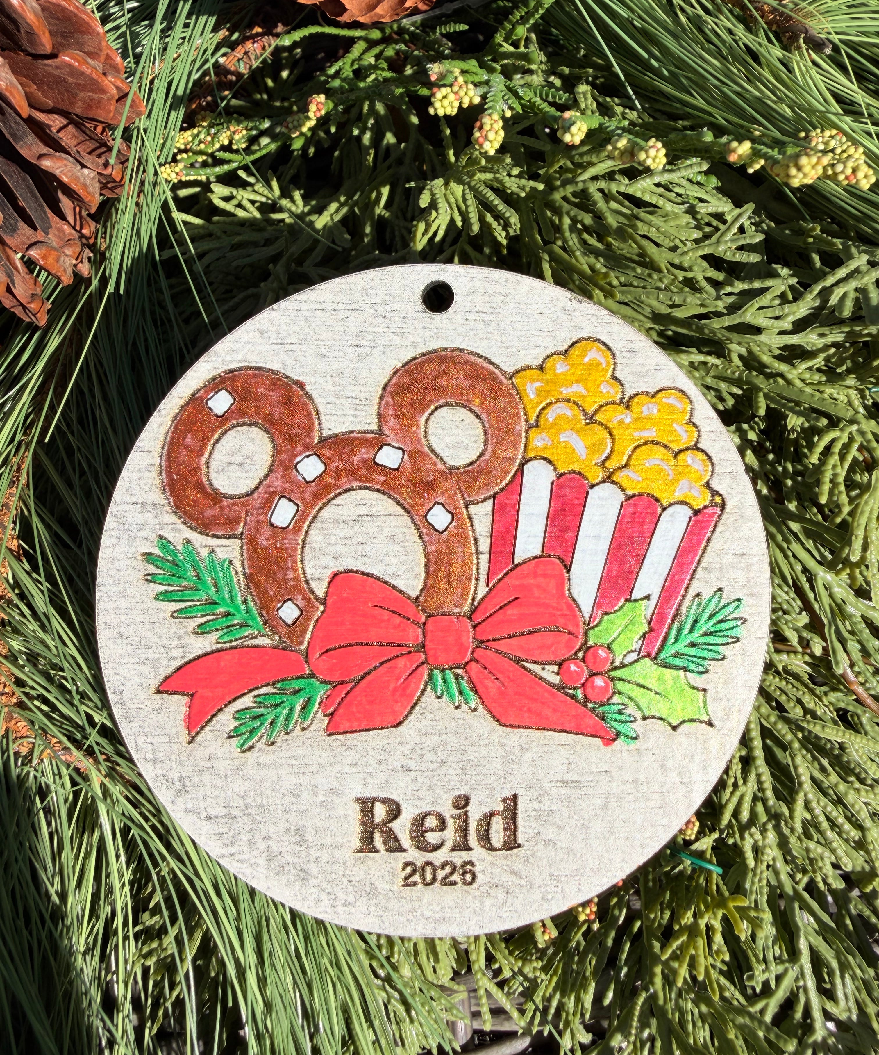 Finished hand-painted Pretzel & Popcorn Christmas Ornament on evergreen greenery. Name and year shown are sample personalization.