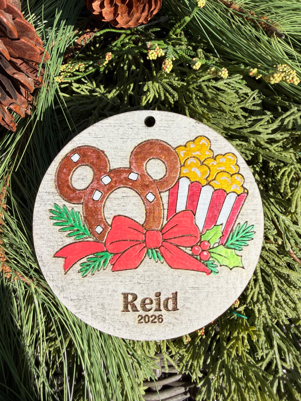 Finished hand-painted Pretzel & Popcorn Christmas Ornament on evergreen greenery. Name and year shown are sample personalization.