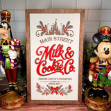 Vertical framed Christmas sign with red Milk & Cookie Co. lettering, castle and evergreen artwork, a cream background and brown wood frame.