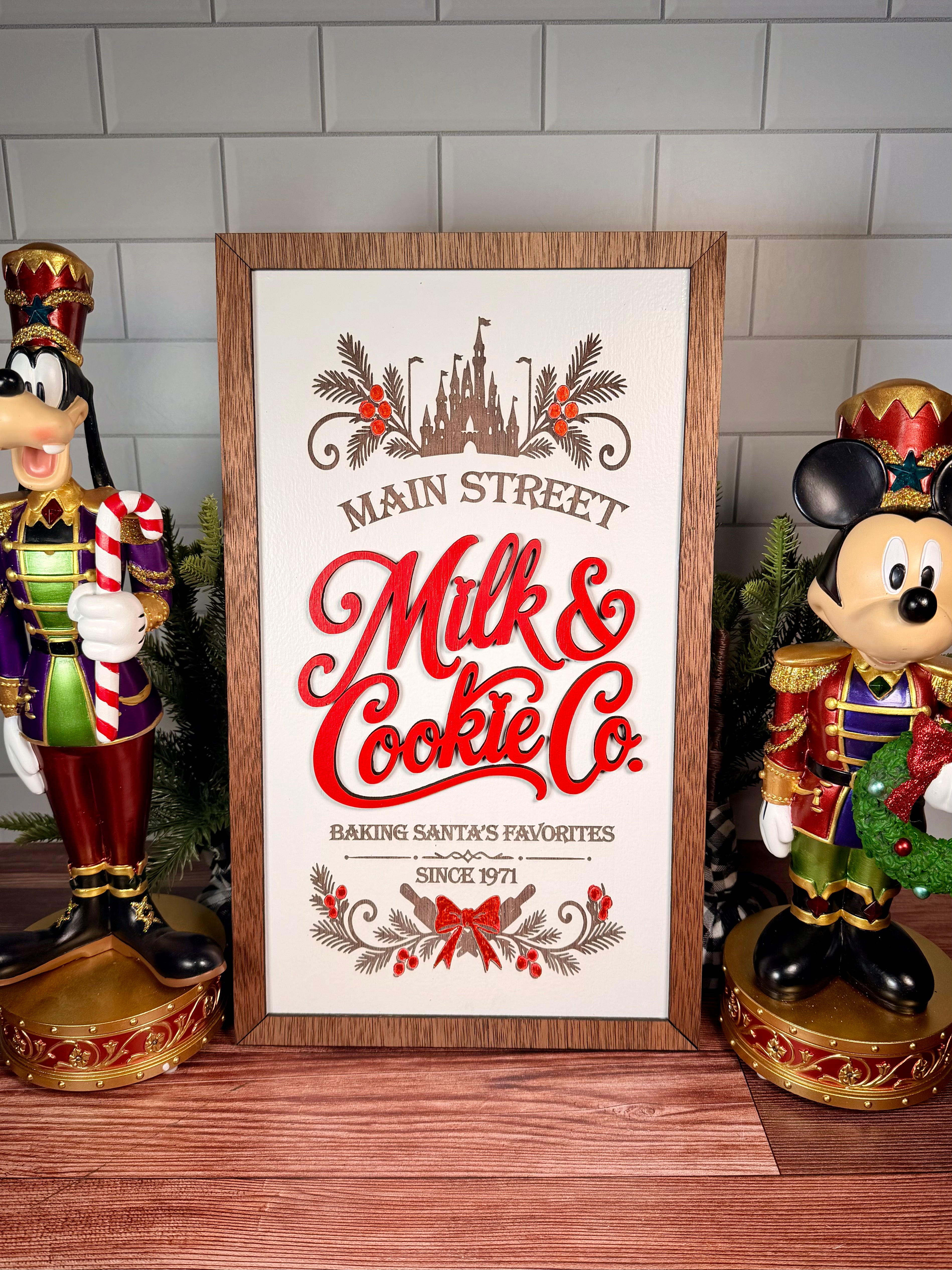 Vertical framed Christmas sign with red Milk & Cookie Co. lettering, castle and evergreen artwork, a cream background and brown wood frame.