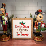 Santa Claus Is Coming to Town square Christmas sign with red lettering, green holly and a brown wood frame.