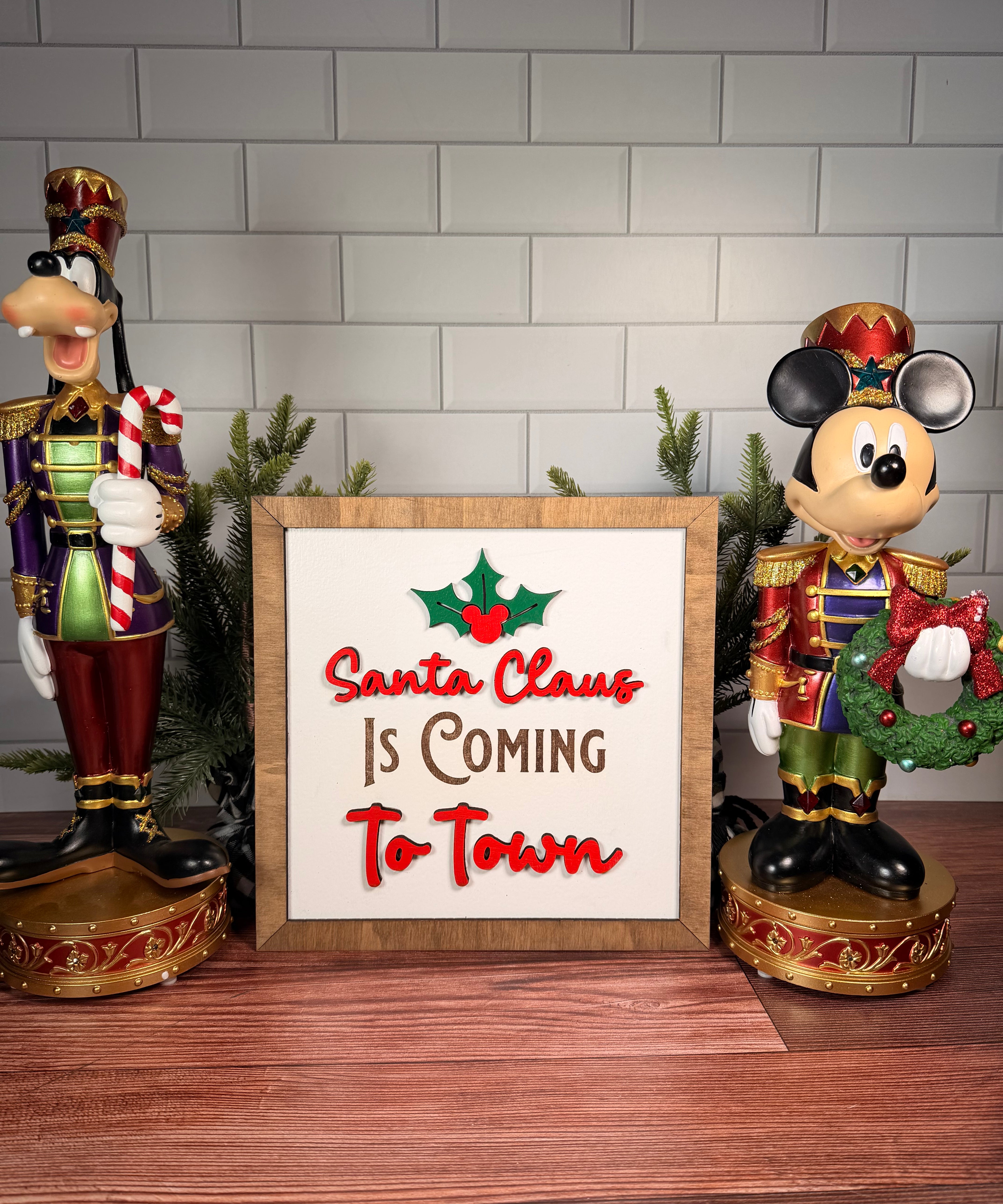 Santa Claus Is Coming to Town square Christmas sign with red lettering, green holly and a brown wood frame.