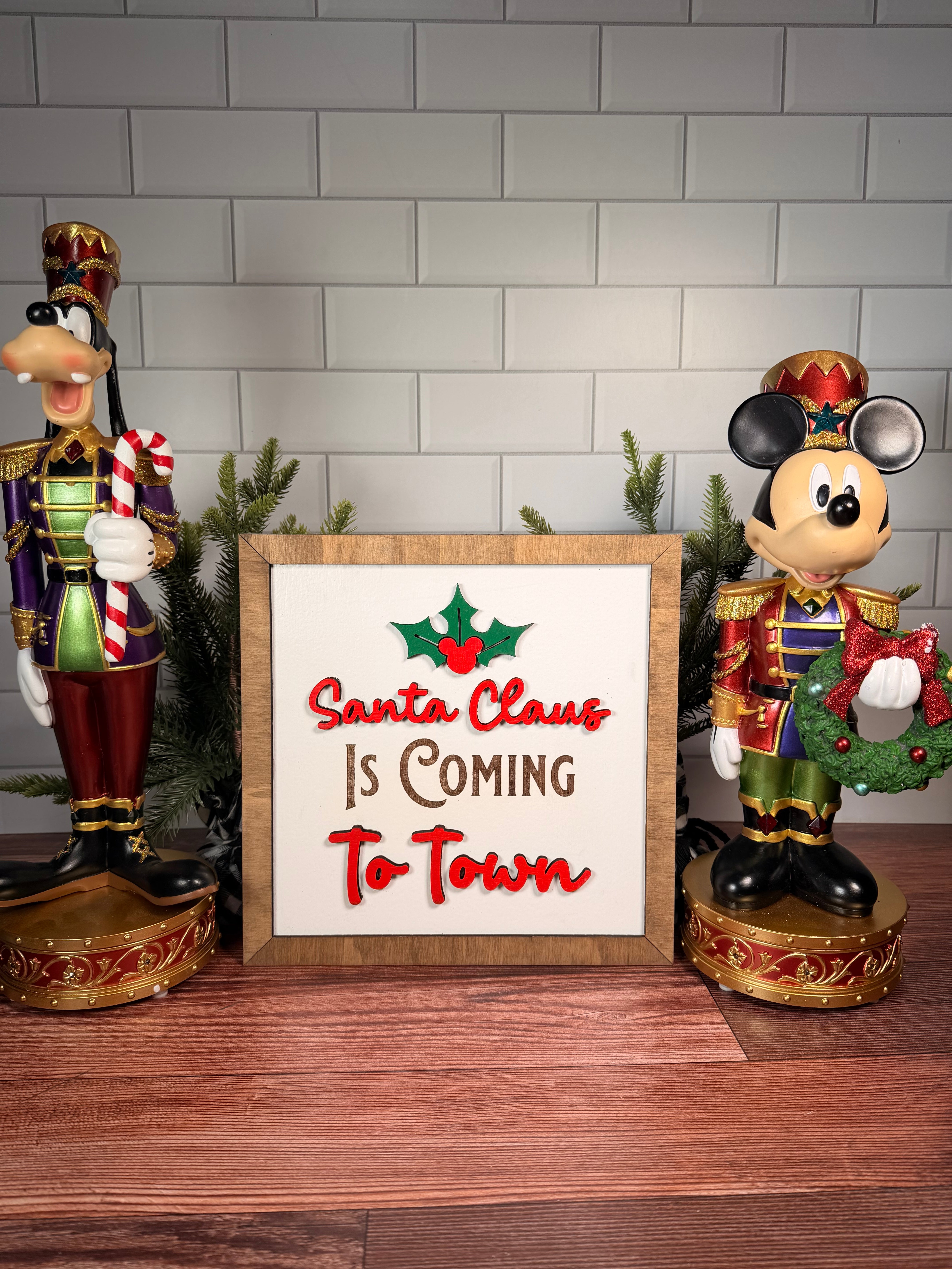 Santa Claus Is Coming to Town square Christmas sign with red lettering, green holly and a brown wood frame.